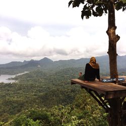 Rear view of woman sitting by tree on wooden platform against mountains