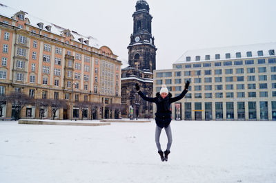 Front view of woman jumping at snow covered town square against sky
