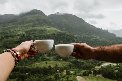 Cropped hands of couple toasting coffee cups against mountain