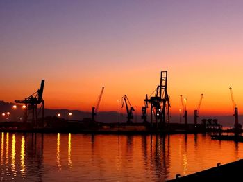 Silhouette cranes at harbor against sky during sunset