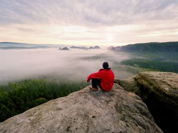 Enjoy life. man in black trousers, jacket and dark cap sit on cliff's edge and looking to mist