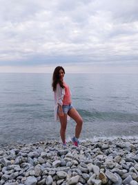 Full length of young woman standing on stones at beach against sky