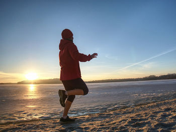 Regular winter workout. runner run by frozen river on sandy beach. orange sun close hilly horizon