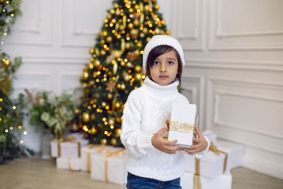 Boy in a white knitted sweater and hat stands with gift box  christmas tree at home on christmas day