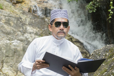 Mature man reading book against waterfall in forest