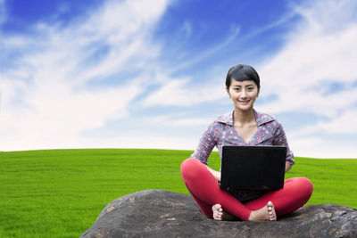 Portrait of smiling woman with laptop sitting against sky