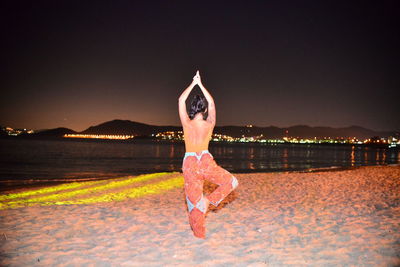 Rear view of mid adult woman exercising at beach against sky during night