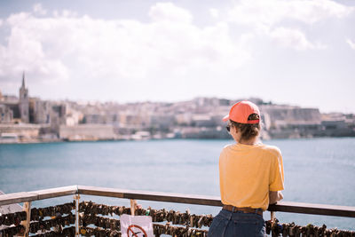 Rear view of young woman looking at river while standing by railing against sky