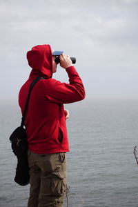 Side view of man looking through binocular while standing against sky