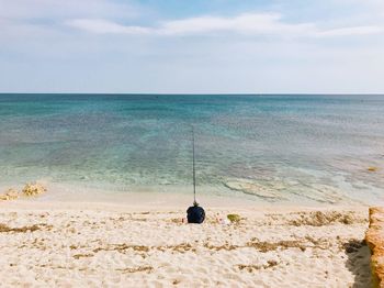 Rear view of man fishing while sitting on shore at beach against sky