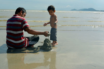 Father and son playing with sand on shore at beach during sunny day