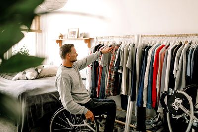 Mature man sitting on wheelchair while choosing clothes from rack at home