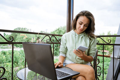 Young woman sits on a balcony at a table working with a phone behind a laptop