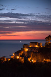 Illuminated building by sea against sky at sunset