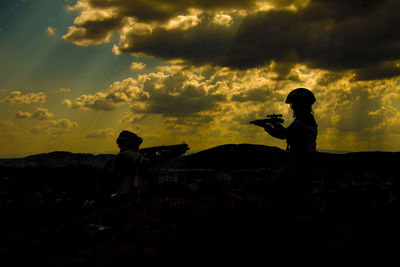Silhouette people paintballing against cloudy sky during sunset