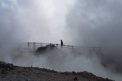 Man walking on a wooden bridge through a thick smoke on top of the crater lake