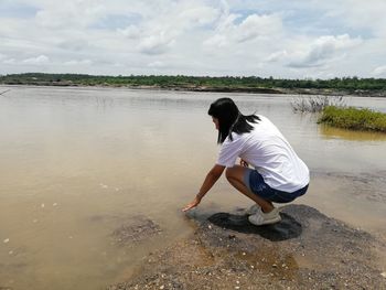 Woman crouching by river against sky