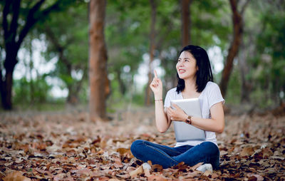 Smiling woman holding digital tablet while sitting on autumn leaves in park