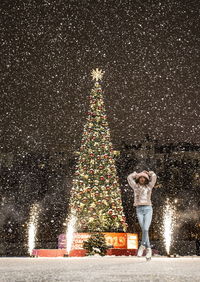 Full length of woman standing by illuminated christmas tree during winter