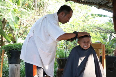 Mature barber trimming scared boy hair at shop against trees