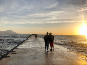 Rear view of people standing on beach against sky during sunset