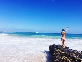 Rear view of woman standing with arms raised on rock formation at beach against blue sky