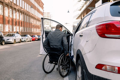 Rear view of mature man boarding white car while sitting on wheelchair in city