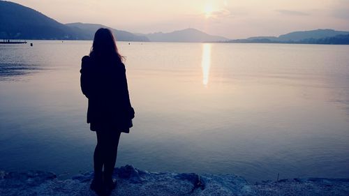 Rear view of woman standing next to lake against sky during sunset