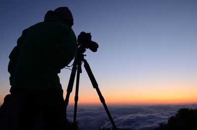 Silhouette person photographing on cliff against sky during sunset