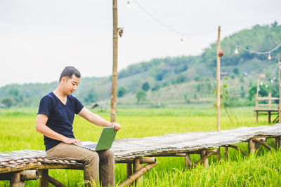 Man using laptop while sitting on boardwalk at rice paddy