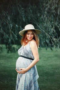 Smiling pregnant woman touching abdomen while standing on grass