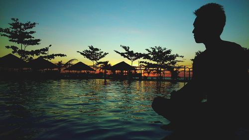 Silhouette man sitting by infinity pool against clear sky during sunset