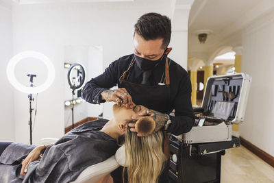 Man in mask smearing foundation of face of blond woman during work in professional makeup studio