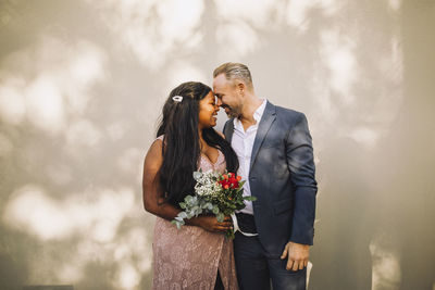 Affectionate young bride holding bouquet standing face to face with groom against wall