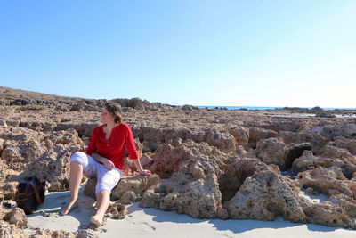 Woman sitting on rocky beach against clear sky