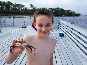 Portrait of shirtless happy boy holding crab while standing on pier