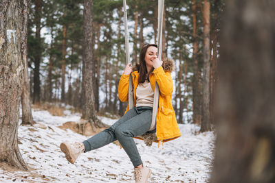 Young carefree woman in yellow jacket swings on swing in winter forest, unity with nature