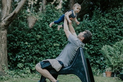 A young handsome dad throws up his daughter while sitting on a garden chair.