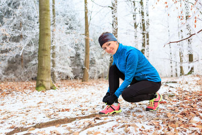 Portrait of mid adult woman tying shoelace while crouching on snow covered field in forest