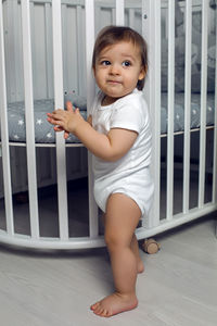 Child in white clothes of one year stands on the floor next to his round white bed