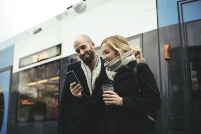 Smiling business people using mobile phone while standing against tram