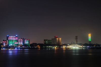 Illuminated buildings by river against sky at night