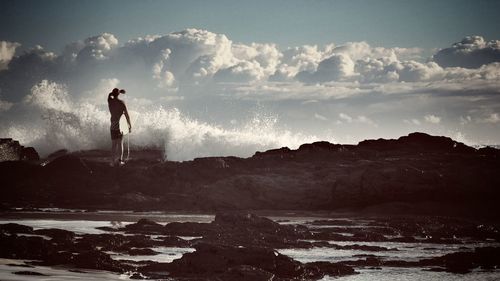 Rear view of man standing with surfboard at rocky sea shore