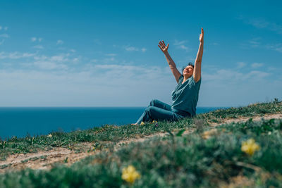 An adult woman sits on a mountain barefoot, raising her hands to the sky and the sun on a sunny day