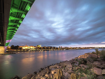 Illuminated buildings by river against cloudy sky