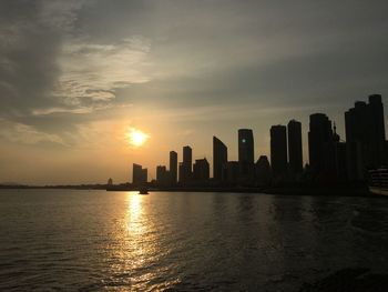 Sea by silhouette buildings against sky during sunset