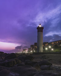 Lighthouse by illuminated buildings against sky at dusk