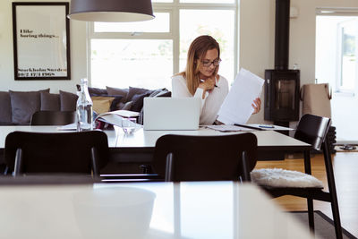 Serious mid adult woman using laptop while reading bills at table in living room