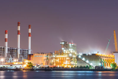 Illuminated factory by river against sky at night