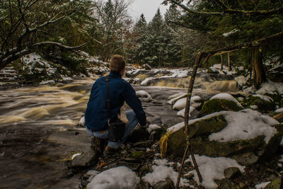 Rear view of man crouching by river in forest during winter